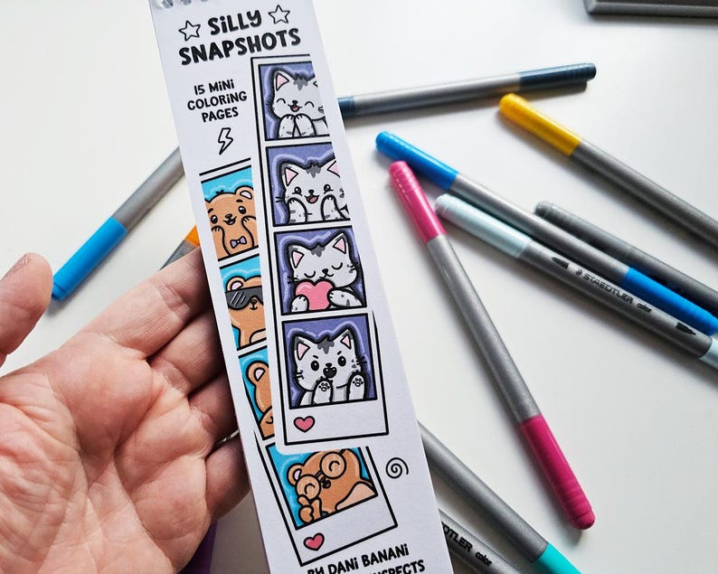 May include: A hand holds a bookmark with the text "Silly Snapshots" and "15 Mini Coloring Pages." The bookmark features cartoon cats and bears in polaroid-style frames. Several gray and silver colored markers with different colored tips are scattered around the bookmark.