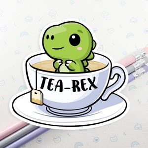 May include: A sticker featuring a green cartoon dinosaur in a teacup. The teacup is white with the words "TEA-REX" in black. The dinosaur has a friendly expression. The teacup sits on a white saucer. Pencils are in the background.
