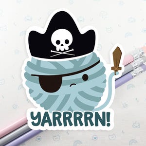 May include: A blue yarn ball wearing a black pirate hat with a skull and crossbones design. The yarn ball has a black eye patch and is holding a sword. The text "YARRRRRN!" is below the yarn ball.