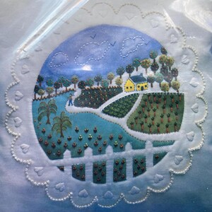 May include: A white fabric with a hand-stitched design of a small town with a yellow house, green trees, and a blue river. The design is surrounded by a white lace border.