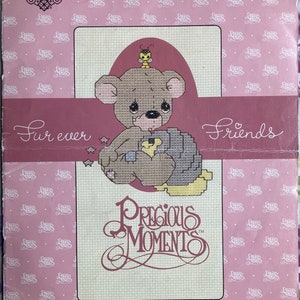 May include: Cross stitch pattern featuring a brown teddy bear holding a yellow chick with a bee on top of the bear's head. The pattern is titled "Fur ever Friends" and includes the Precious Moments logo.