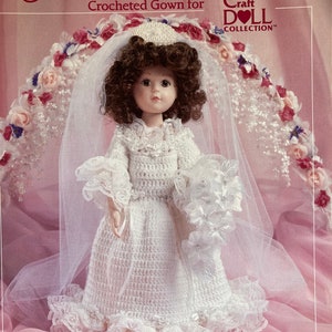 May include: Crocheted wedding gown for a 14-inch doll. The gown is white with a lace trim and a veil. The doll is wearing a white headband with pearls. The doll is standing in front of a pink background with a floral design.