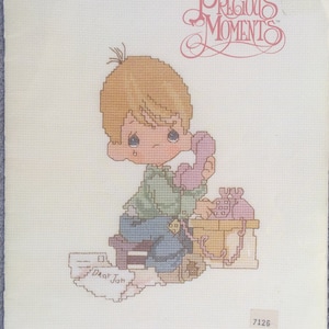 May include: Cross stitch pattern featuring a Precious Moments character, a young boy with blonde hair, sitting on a chair and talking on a pink phone. The pattern is titled "Dear Jon" and is from book PM-3.