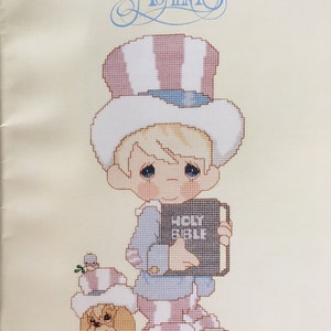 May include: Cross-stitch pattern featuring a Precious Moments character dressed as Uncle Sam holding a book that says "HOLY BIBLE". The character is wearing a red, white, and blue striped outfit and a hat with stars. A small dog is beside the character.