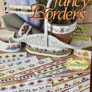 Puede incluir: Un libro titulado "Fancy Borders" con una cesta de mimbre blanca y varios dise&ntilde;os de punto de cruz para ribetes y decoraci&oacute;n. El libro es de la tienda NeedleCraft Shop y presenta 21 dise&ntilde;os de punto de cruz f&aacute;ciles de hacer.