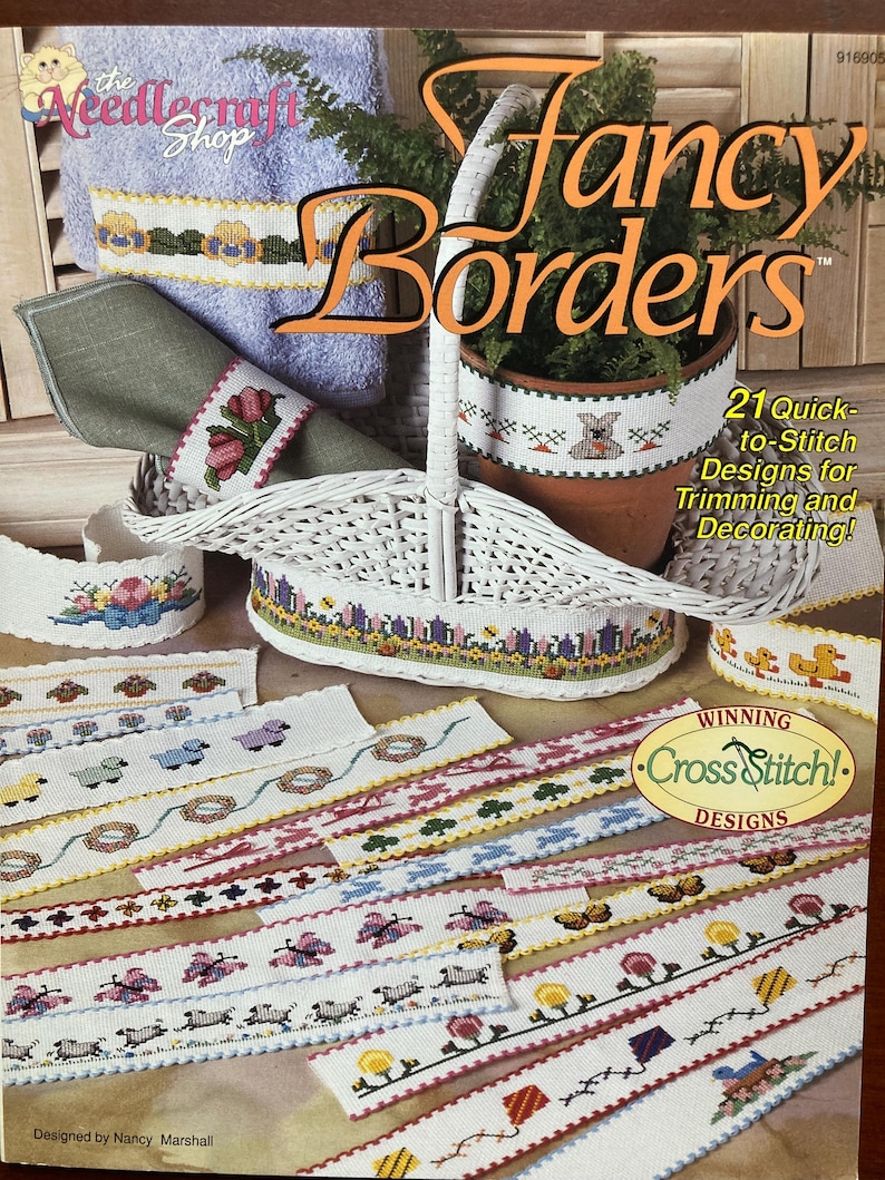 Puede incluir: Un libro titulado "Fancy Borders" con una cesta de mimbre blanca y varios dise&ntilde;os de punto de cruz para ribetes y decoraci&oacute;n. El libro es de la tienda NeedleCraft Shop y presenta 21 dise&ntilde;os de punto de cruz f&aacute;ciles de hacer.
