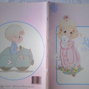 May include: A cross stitch pattern booklet featuring two Precious Moments characters. The first image shows a boy in a blue and white checkered shirt driving a green car. The second image shows a girl in a pink dress holding a white lamb. The text "Precious Moments" is printed on the cover.