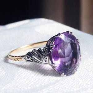 Può includere: Un anello in stile vintage con una grande pietra preziosa viola taglio ovale. L'anello ha una fascia d'oro con dettagli argentati intorno alla gemma. Poggia su una superficie bianca strutturata.