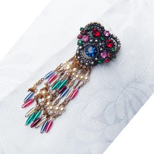 Puede incluir: Broche vintage con diseño de corazón, adornado con piedras preciosas rojas, rosas, verdes y azules. El broche presenta una piedra azul central y hebras colgantes de cuentas de varios colores. Fondo blanco.