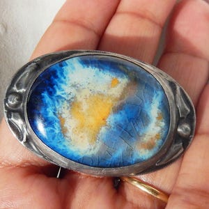 May include: Oval-shaped silver brooch with a vibrant, abstract design. The center features a cracked, glazed surface with shades of blue, yellow, and white. The silver frame has decorative detailing around the edges. A unique piece of jewelry.