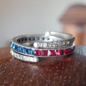 May include: A silver ring with three bands, each adorned with gemstones. One band features blue stones, another red, and the third clear. The ring is set on a wooden surface.