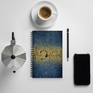 May include: A blue spiral notebook with a gold glitter design and a black heartbeat graphic with a headset and a heart. The notebook is on a white surface with a coffee cup, a pencil, and a black smartphone.