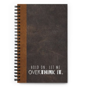 May include: A brown leather-bound spiral notebook with a brown spine. The cover has the text "HOLD ON. LET ME OVERTHINK IT."