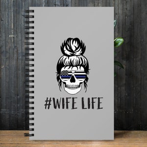 Puede incluir: Cuaderno gris de espiral con un gráfico de calavera en blanco y negro que lleva gafas de sol con una bandera estadounidense y una fina línea azul. El texto #WIFE LIFE está impreso debajo de la calavera.