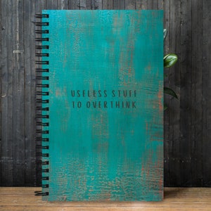 May include: A teal blue spiral bound notebook with a distressed wood-like texture. The cover has the text "Useless Stuff To Overthink" printed in black.