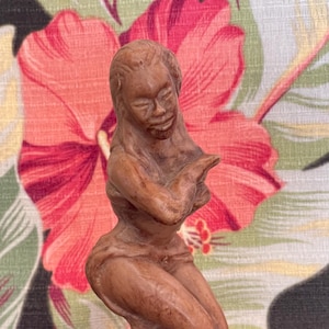 May include: A brown ceramic figurine of a kneeling woman with arms crossed. The figurine is on a round base with the word "HAWAIIAN" inscribed. The background features a floral pattern with red and pink flowers.