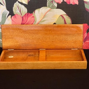 May include: A rectangular wooden box with a hinged lid, featuring a polished light brown finish and a visible wood grain. The interior is divided into two separate open compartments, with a small circular sticker visible in the left section.