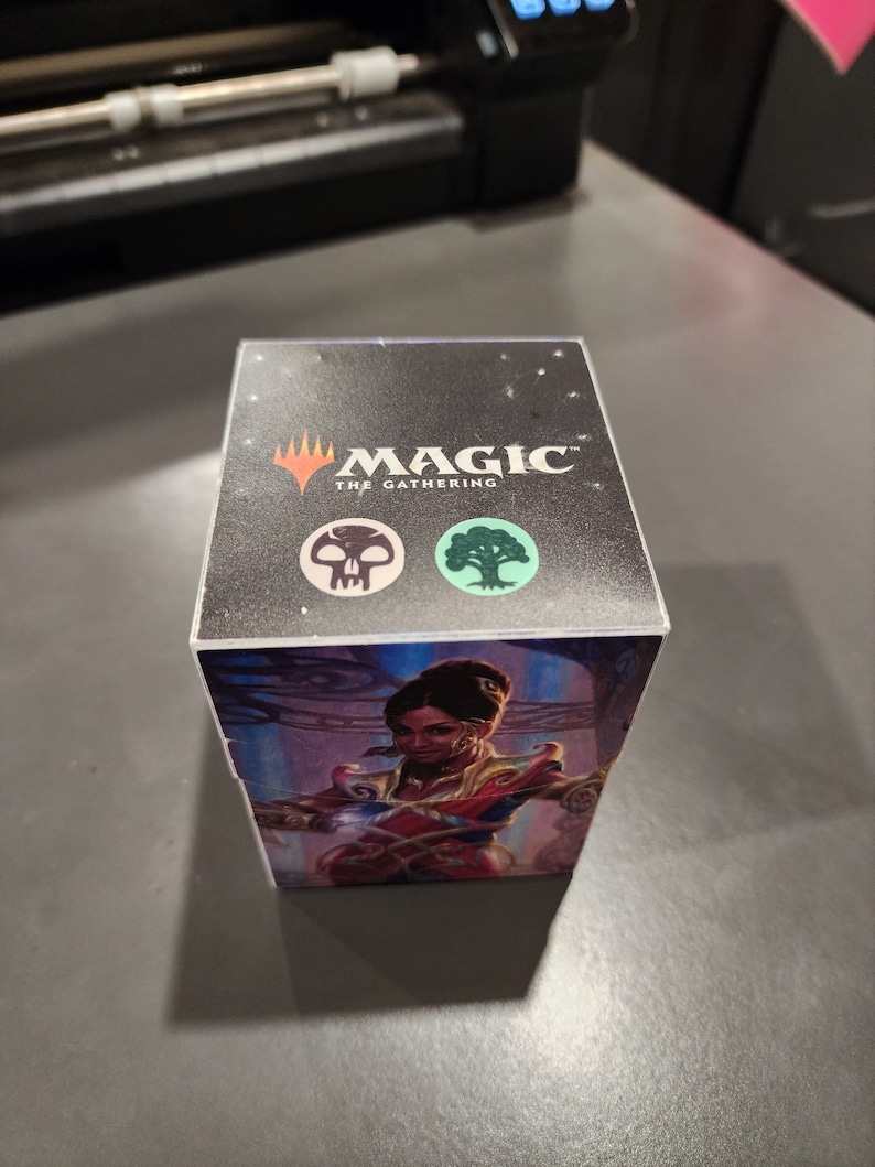 May include: A black Magic: The Gathering deck box with a woman in a red and gold outfit on the front. The box has a black skull and a green tree symbol on the top.