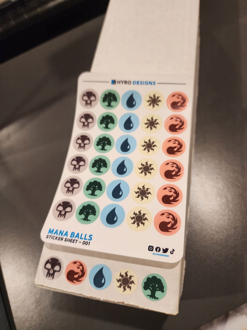 May include: A sticker sheet with 36 colorful mana ball stickers. The sheet features a variety of symbols, including skulls, trees, drops of water, suns, and flames. The sheet is labeled "Mana Balls Sticker Sheet - 001".