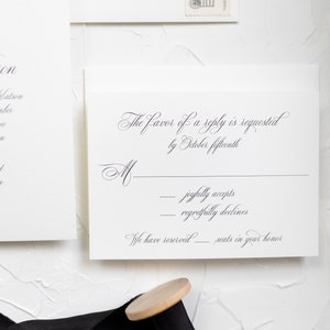 May include: A white RSVP card with black text. The text reads "The favor of a reply is requested by October fifteenth."  There are two lines for the guest to check off: "joyfully accepts" and "regretfully declines". The card also states "We have reserved ______ seats in your honor."