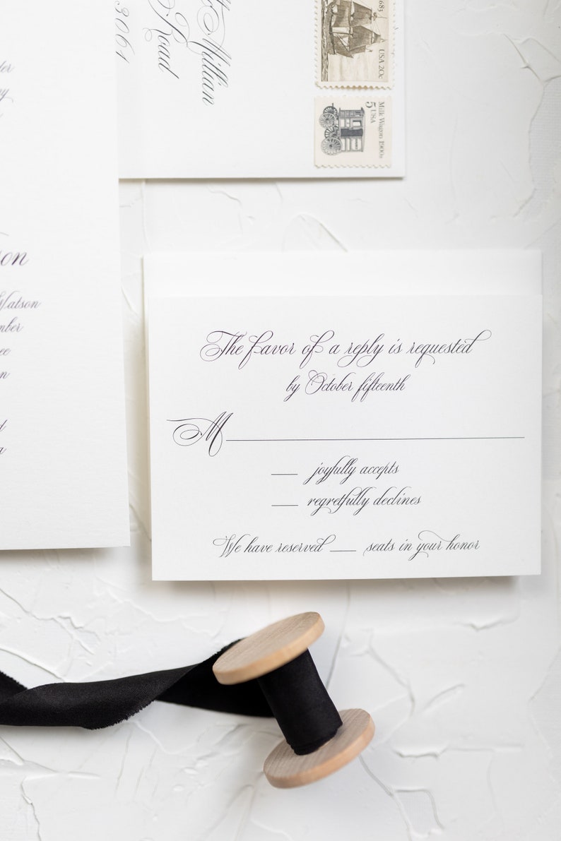 May include: A white RSVP card with black text. The text reads "The favor of a reply is requested by October fifteenth."  There are two lines for the guest to check off: "joyfully accepts" and "regretfully declines". The card also states "We have reserved ______ seats in your honor."