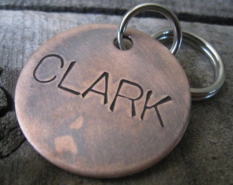 May include: A round copper dog tag with the name "CLARK" engraved on it. The tag is attached to a silver split ring.