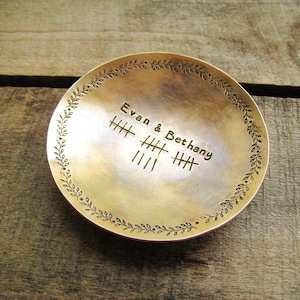 May include: A small, round, brass-colored dish with the names "Evan & Bethany" and tally marks etched into the center. The rim is decorated with a delicate leaf pattern. The dish sits on a wooden surface.