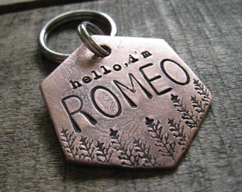 May include: Copper dog tag with a hexagonal shape and a key ring. The tag is engraved with the text "hello, i'm ROMEO" and a decorative design of leaves.