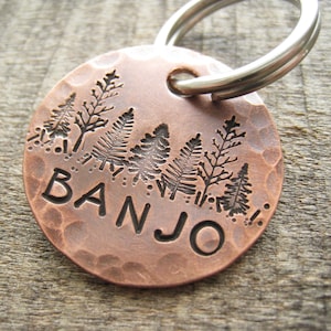 May include: A copper dog tag with a hammered finish and a design of stylized trees. The tag is engraved with the name "BANJO".