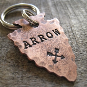 May include: A copper arrowhead keychain with the word "ARROW" engraved on it. The arrowhead has a hammered texture and features a small cross-shaped arrow design.