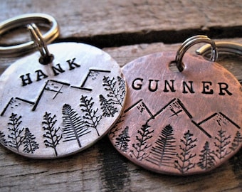 May include: Two silver and copper dog tags with engraved mountain and tree designs. The tags read "Hank" and "Gunner".