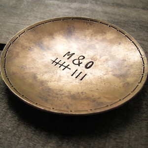 May include: A round, antique brass dish with a textured surface. The dish features the stamped text "M & O" above a series of tally marks and the number "III". The rim is decorated with small, evenly spaced dots.