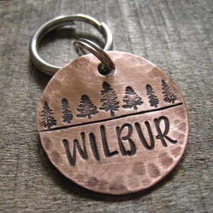 May include: Copper dog tag with a hammered finish, featuring a row of pine trees and the name "WILBUR" engraved in a cursive font.