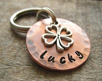 May include: A copper dog tag with a hammered finish and a silver four-leaf clover charm. The tag is engraved with the word "lucky".