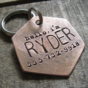 May include: Copper dog tag with a hexagonal shape and a silver key ring. The tag is engraved with the text "hello, i'm RYDER 555.732.9918".