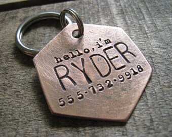 May include: Copper dog tag with a hexagonal shape and a silver key ring. The tag is engraved with the text "hello, i'm RYDER 555.732.9918".