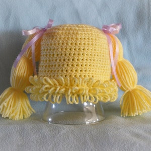 May include: A bright yellow crocheted hat featuring a textured loop-stitch brim and two attached yarn pigtails on either side. Each pigtail is secured with a light pink satin ribbon tied into a bow, adding a decorative accent to the yarn hair.