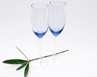 Puede incluir: Dos copas de champán de vidrio transparente con bordes azules. Las copas están sobre una superficie blanca con una ramita verde delante de ellas.