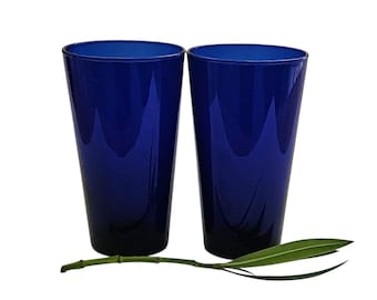 Può includere: Due bicchieri cilindrici alti realizzati in vetro blu cobalto intenso. I bicchieri presentano un design liscio e affusolato con una finitura lucida. Un singolo stelo di pianta verde con foglie lunghe e sottili giace orizzontalmente davanti ai bicchieri.