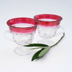 Könnte beinhalten: Zwei klare Glasbecher mit rotem Rand und einem klaren Glasfuß. Die Becher haben ein gewelltes Design und stehen auf einer weißen Oberfläche.