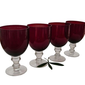 May include: A set of four wine glasses featuring deep red, cranberry-coloured bowls and clear glass stems with a rounded, tiered base design. The glasses are arranged in a row, showcasing the contrast between the dark red glass and the clear base.