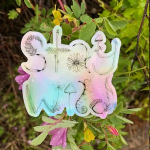 May include: A holographic sticker with the words "Stay Wild" written in a whimsical font. The sticker features a variety of botanical illustrations, including mushrooms, ferns, and flowers.