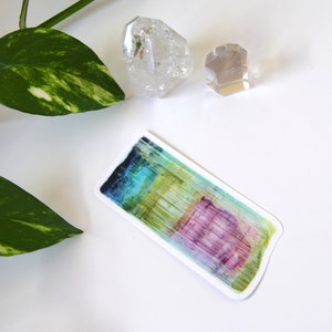 May include: A colorful sticker with a watercolor-like design of a crystal. The sticker is rectangular and has a white border. The crystal is a mix of blue, green, yellow, and pink.