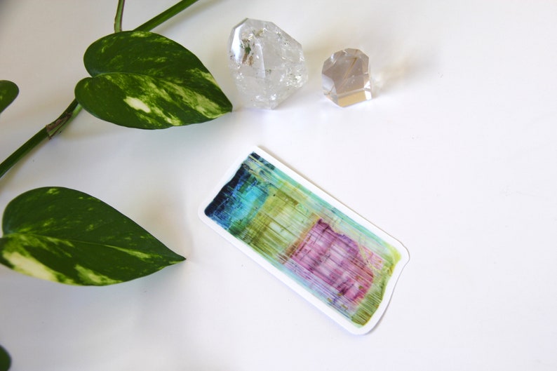 May include: A colorful sticker with a watercolor-like design of a crystal. The sticker is rectangular and has a white border. The crystal is a mix of blue, green, yellow, and pink.