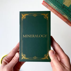 May include: A dark green book titled "MINERALOGY" with gold ornate border and lettering. The book is held by two hands. Two antique books are in the background. A magnifying glass is in the lower left corner.