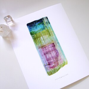 May include: A close-up of a colorful, striated tourmaline crystal. The crystal is a vibrant mix of green, blue, purple, and yellow. The crystal is on a white background with the word "TOURMALINE" printed below it.