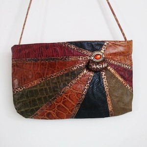 May include: A rectangular shoulder bag with a patchwork design in shades of brown, red, and black. The bag features a central decorative clasp with an orange stone. The strap is a thin, brown cord.