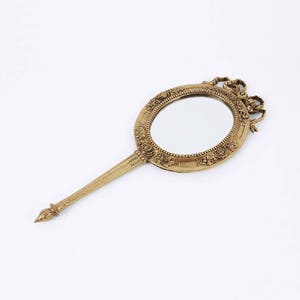 May include: A gold-toned hand mirror with an ornate, oval-shaped frame featuring floral and ribbon details. The mirror has a long, slender handle.