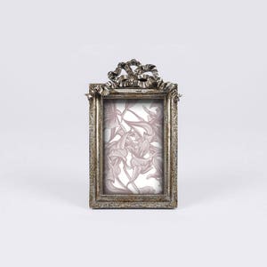May include: A silver-toned picture frame with a decorative bow design on the top. The frame has a floral pattern inside.