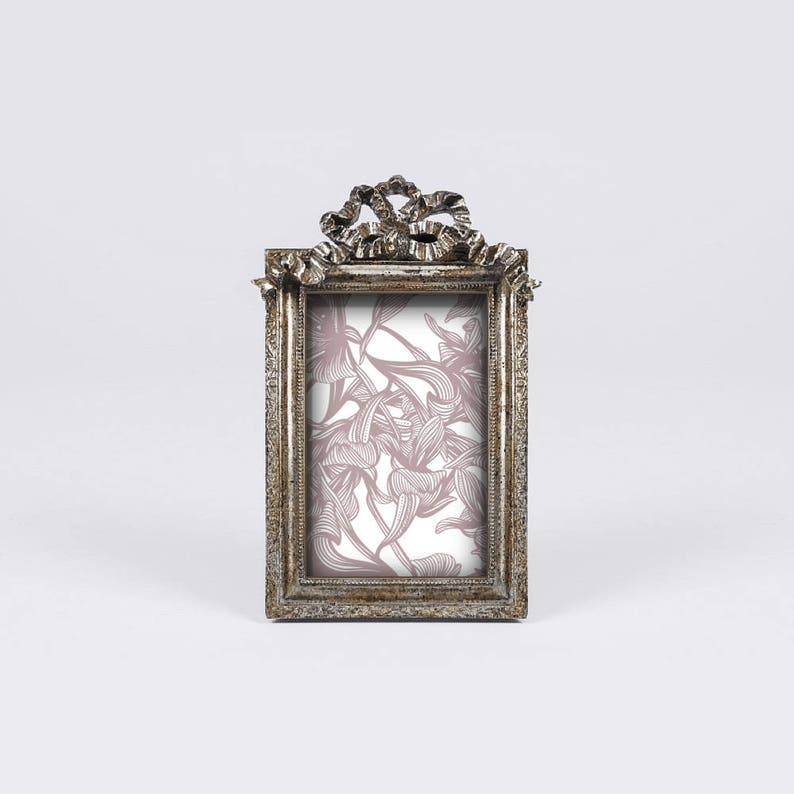 May include: A silver-toned picture frame with a decorative bow design on the top. The frame has a floral pattern inside.